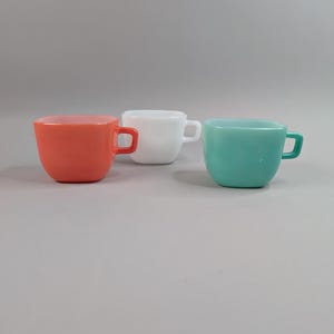 Peut inclure: Trois tasses à café carrées, de couleurs corail, blanche et turquoise. Chaque tasse est dotée d'une petite anse carrée. Les tasses sont en matériau brillant et sont disposées sur un fond gris uni. Idéales pour le café ou le thé.