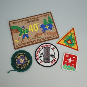 Peut inclure: Cinq écussons brodés, dont un écusson brun avec le texte "TRI-DISTRICT PAUL BUNYAN'S CAMPOREE 40th 1998 ROBERT MOSES STATE PARK N.Y.", un écusson jaune triangulaire avec le texte "TREES FOR CANADA EST 1966", un écusson rond vert avec le texte "DISTRICT LAC SAINT-JEAN", un écusson rond blanc avec le texte "SYDENHAM DISTRICT 92 SINISH DERBY", et un écusson carré rouge avec le texte "DISTRICT 0 HURON".