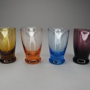 May include: Four small glass shot glasses in different colours. The glasses are clear with coloured bottoms. From left to right, the colours are amber, orange, blue, and purple.