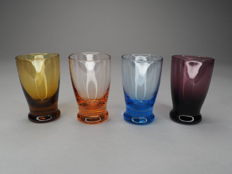 May include: Four small glass shot glasses in different colours. The glasses are clear with coloured bottoms. From left to right, the colours are amber, orange, blue, and purple.