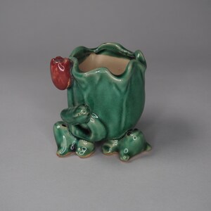 May include: A green ceramic planter shaped like a frog with a red flower bud on its back. The planter has four legs and a wide opening at the top.