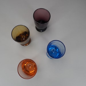 May include: Four empty glass shot glasses in different colours: purple, brown, blue, and orange. The glasses are on a white surface.