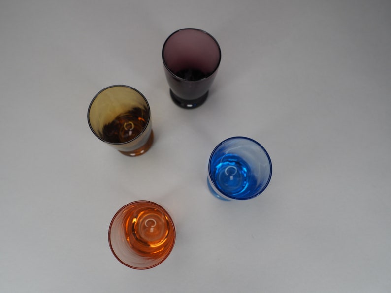 May include: Four empty glass shot glasses in different colours: purple, brown, blue, and orange. The glasses are on a white surface.