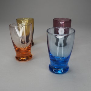 May include: Four clear glass shot glasses with coloured bases. The glasses are arranged in a row, with the bases in the following order: orange, yellow, purple, and blue.