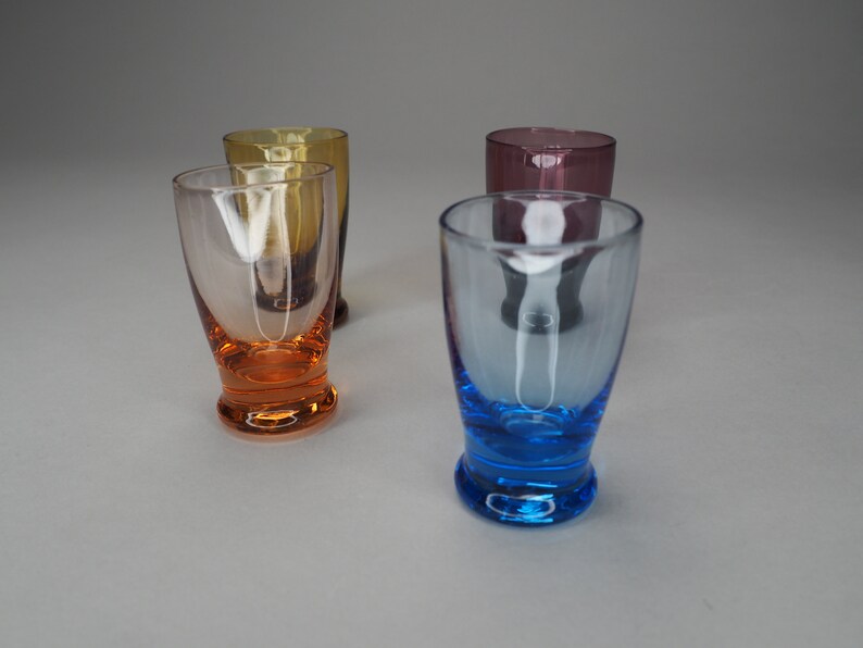 May include: Four clear glass shot glasses with coloured bases. The glasses are arranged in a row, with the bases in the following order: orange, yellow, purple, and blue.