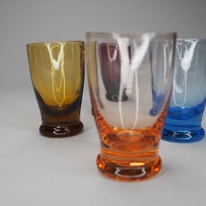 May include: Four small glass shot glasses in different colours. The glasses are clear, amber, blue, and orange.