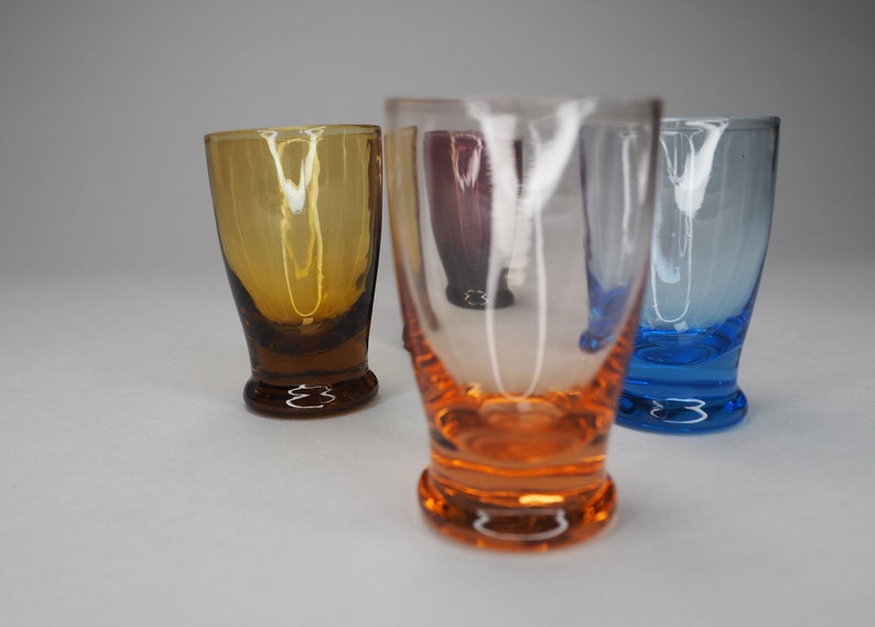 May include: Four small glass shot glasses in different colours. The glasses are clear, amber, blue, and orange.