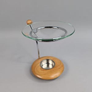May include: An aromatherapy oil warmer with a clear glass dish, chrome metal accents, and a wooden base. The base has a circular design with a silver metal cup for a tea light. The warmer is designed to hold essential oils.
