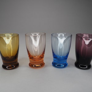 May include: Four shot glasses in different colours: amber, orange, blue, and purple. The glasses are all clear and have a rounded bottom.
