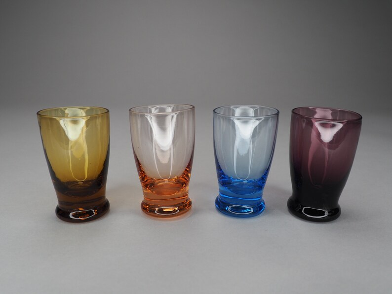 May include: Four shot glasses in different colours: amber, orange, blue, and purple. The glasses are all clear and have a rounded bottom.
