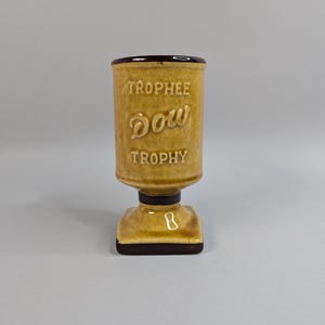 May include: A tan ceramic trophy with the word "TROPHEE" above "Dow" and "TROPHY" in embossed lettering. The trophy has a dark brown rim and base, and a square base with a small cutout.