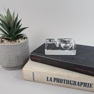 May include: A clear glass candle holder with two separate compartments. The holder is sitting on top of two books, one black and one white. The white book has the text "LA PHOTOGRAPHIE" printed on the cover.