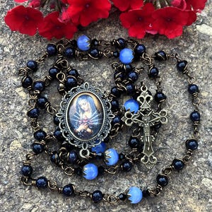 May include: A rosary featuring dark blue glittery beads and light blue smooth beads connected by bronze-toned wire. The centerpiece is an oval pendant depicting a figure with rays of light, and the crucifix is an ornate bronze-toned metal cross.
