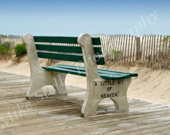 May include: A green bench with a white inscription that reads "A little bit of heaven" sits on a wooden boardwalk. The bench is facing away from the camera and is positioned in front of a wooden fence and sand dunes.