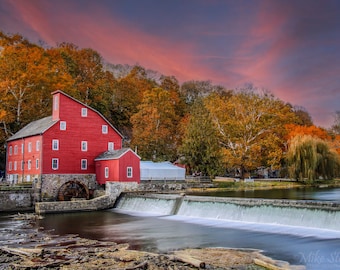 May include: A red wooden watermill with a water wheel sits on a river with a waterfall. The mill is surrounded by fall foliage in shades of orange, yellow, and red. The sky is a soft pink and purple.