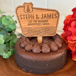 May include: Wooden sign with a rustic design, featuring the text "Steph & James Let the Married Adventures Begin Ouray, CO". The sign is decorated with a pine tree and a shield.