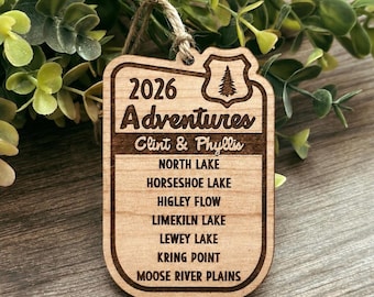 May include: A rectangular wooden ornament with rounded corners and a jute twine loop. The engraving reads 2026 Adventures, Clint & Phyllis, and a list of locations: North Lake, Horseshoe Lake, Higley Flow, Limekiln Lake, Lewey Lake, Kring Point, Moose River Plains.