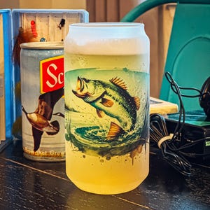 May include: A frosted glass beer can-shaped with a jumping bass fish graphic. The glass is filled with a light-colored liquid, possibly beer, and has a white foam head. A vintage fishing tackle box and a can with a duck graphic are in the background.