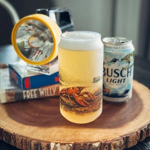 May include: A clear glass can-shaped tumbler filled with a light-colored beverage and topped with white foam. The glass features a detailed graphic of a crawfish on a riverbank. Next to it sits a Busch Light beer can and a yellow flashlight.