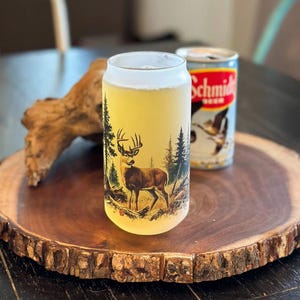 May include: A frosted glass beer can-shaped tumbler with a deer and forest scene design. The glass is filled with beer and sits next to a can of Schmidt beer on a wooden serving tray.