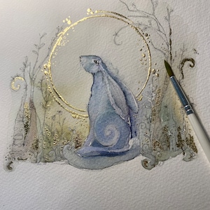 May include: Watercolor painting of a blue hare within a gold circle, surrounded by delicate foliage. The artwork features gold accents and a soft, ethereal quality. A paintbrush is visible in the right corner, suggesting the artist's process.
