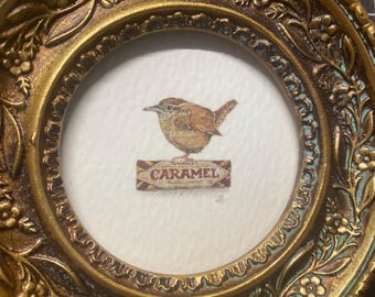 Könnte beinhalten: Ein kleiner, runder, antikgoldfarbener Rahmen mit detailliertem floralem Design. Im Inneren befindet sich ein Aquarell eines braunen Vogels, der über einem Schild mit der Aufschrift "Caramel" sitzt. Das Kunstwerk befindet sich auf weißem Hintergrund.