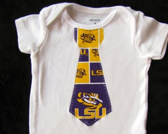 May include: White baby onesie with a purple and gold tie applique featuring the LSU logo and mascot. The onesie has the text "carter's" on the tag.