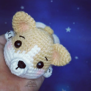 May include: A crocheted stuffed dog toy with brown and white fur, large black eyes, and a black nose. The toy is lying on its back with its paws in the air.