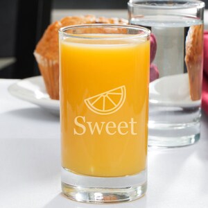 May include: A clear glass with a slice of orange and the word "Sweet" etched on the side, filled with orange juice.