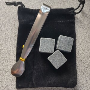 May include: A set of three gray stone whiskey chilling cubes in a black velvet pouch with a silver and gold metal tong.