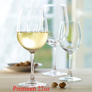 Op de afbeelding: Drie heldere glazen wijnglazen, waarvan één met de naam "Arden" gegraveerd aan de zijkant, gevuld met witte wijn. Het glas met de naam is een premium wijnglas van 22 ounce.