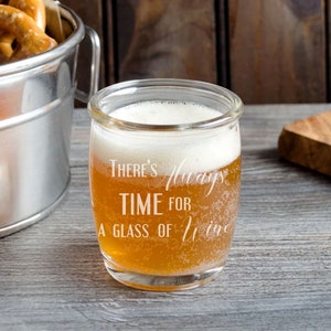 May include: A clear glass tumbler filled with a light amber liquid and white foam. The glass is etched with the words "There's Always Time For A Glass of Wine". A metal bucket with pretzels is in the background.