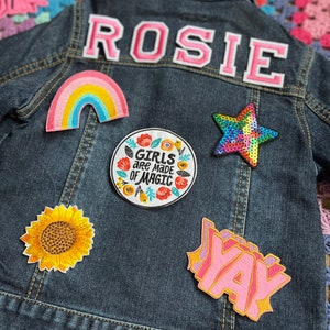 May include: A denim jacket with a variety of colorful patches, including a rainbow, a sunflower, a star, a text patch that says "Girls are made of magic", and a text patch that says "Yay". The jacket also has a pink felt patch with the name "Rosie" on the back.