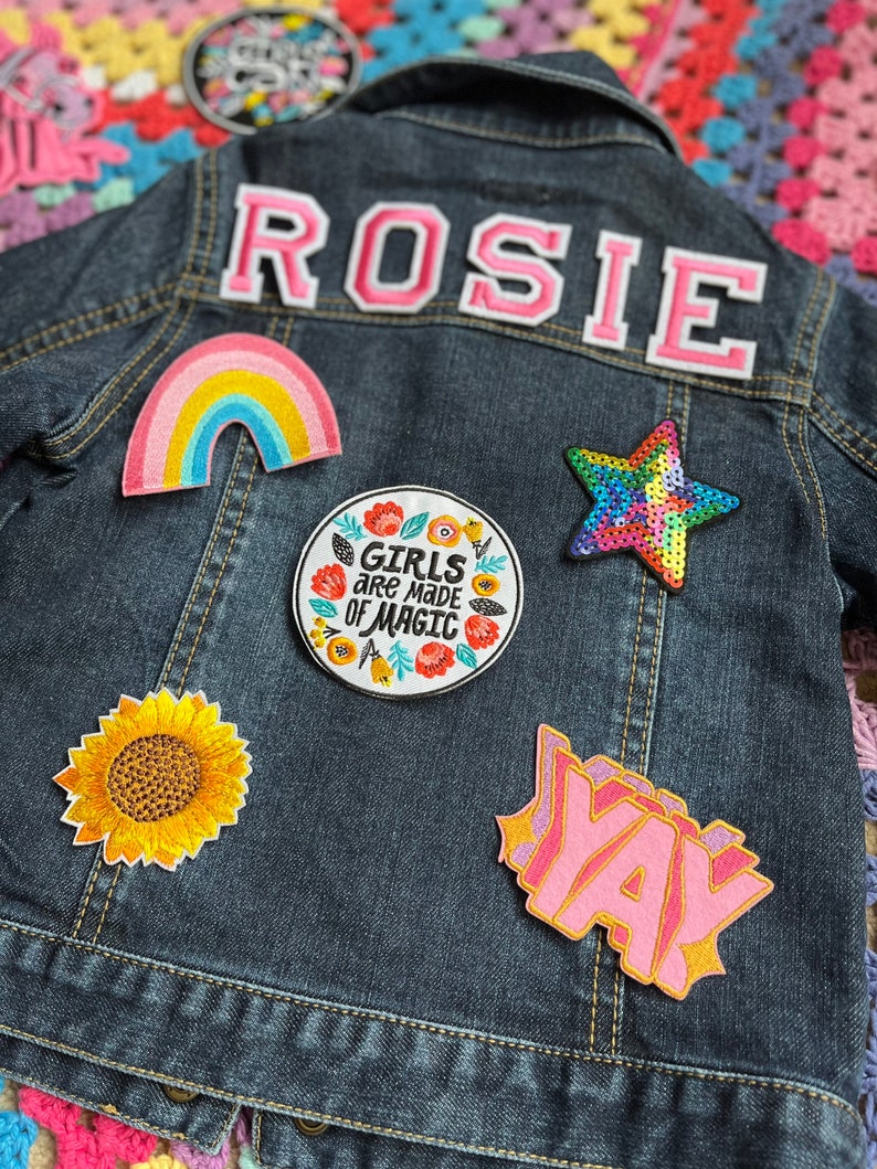 May include: A denim jacket with a variety of colorful patches, including a rainbow, a sunflower, a star, a text patch that says "Girls are made of magic", and a text patch that says "Yay". The jacket also has a pink felt patch with the name "Rosie" on the back.