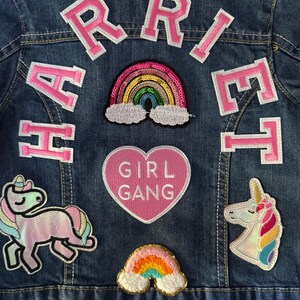 May include: A blue denim jacket with pink felt letters spelling "HARRIET" on the back. The jacket has several patches including a rainbow, a unicorn, a heart with the text "GIRL GANG", and another rainbow.