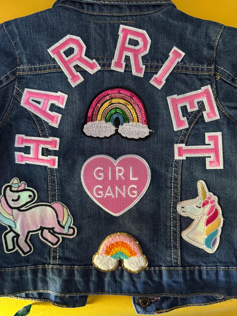 May include: A blue denim jacket with pink felt letters spelling "HARRIET" on the back. The jacket has several patches including a rainbow, a unicorn, a heart with the text "GIRL GANG", and another rainbow.