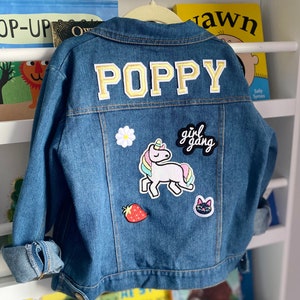 May include: A blue denim jacket with the name "POPPY" embroidered on the back in gold letters. The jacket has several patches, including a unicorn, a strawberry, a cat, and a black patch that says "girl gang".