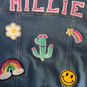 May include: A blue denim jacket with a pink "MILLIE" patch on the back. The jacket has several colorful patches including a rainbow, a cactus, a flower, a smiley face, and a shooting star.