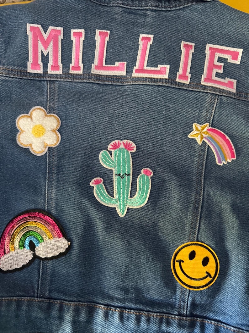 May include: A blue denim jacket with a pink "MILLIE" patch on the back. The jacket has several colorful patches including a rainbow, a cactus, a flower, a smiley face, and a shooting star.