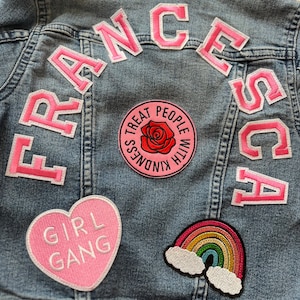May include: A light blue denim jacket with pink patches. The patches spell out "FRANCESCA" in pink letters. Other patches include a heart with the words "GIRL GANG", a rainbow with clouds, and a circular patch with a rose and the words "TREAT PEOPLE WITH KINDNESS".