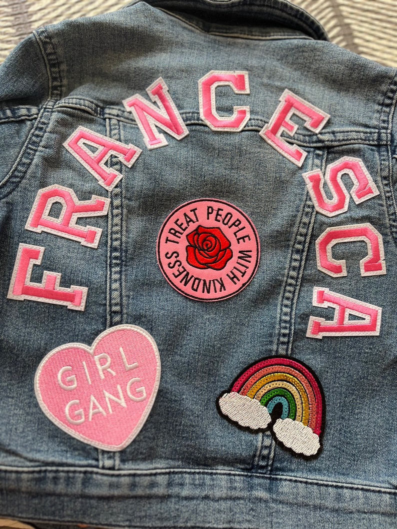 May include: A light blue denim jacket with pink patches. The patches spell out "FRANCESCA" in pink letters. Other patches include a heart with the words "GIRL GANG", a rainbow with clouds, and a circular patch with a rose and the words "TREAT PEOPLE WITH KINDNESS".