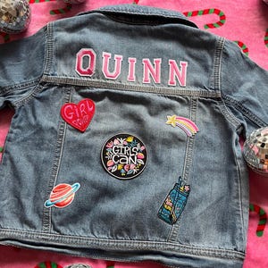 May include: A blue denim jacket with the name "QUINN" embroidered on the back in pink letters. The jacket has several patches, including a pink heart with the words "Girl Power", a black circle with the words "Girls Can" in white, a rainbow shooting star, and a blue mason jar with a rainbow inside.