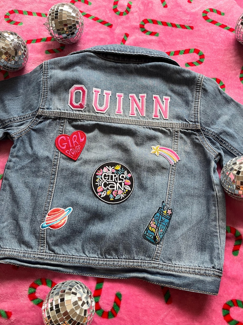 May include: A blue denim jacket with the name "QUINN" embroidered on the back in pink letters. The jacket has several patches, including a pink heart with the words "Girl Power", a black circle with the words "Girls Can" in white, a rainbow shooting star, and a blue mason jar with a rainbow inside.