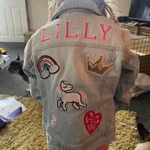 May include: A light blue denim jacket with the name "LILLY" embroidered on the back in pink letters. The jacket has several colorful patches sewn on, including a rainbow, a unicorn, a crown, and a heart with the words "Girl Power" on it.
