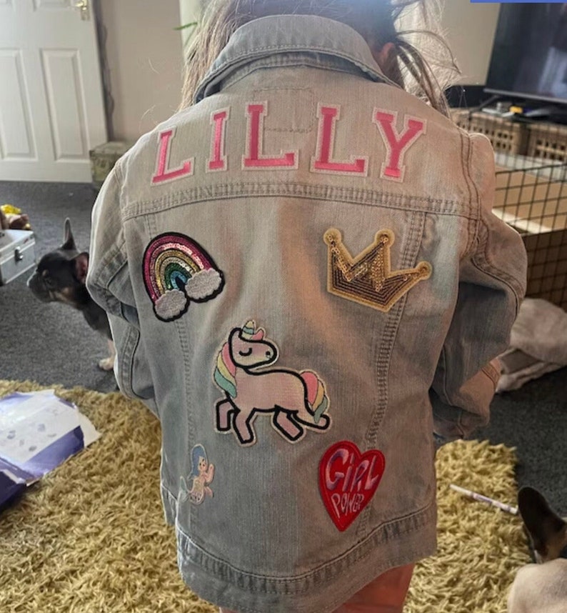 May include: A light blue denim jacket with the name "LILLY" embroidered on the back in pink letters. The jacket has several colorful patches sewn on, including a rainbow, a unicorn, a crown, and a heart with the words "Girl Power" on it.