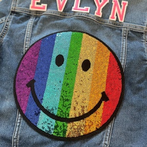 May include: A denim jacket with a rainbow sequin smiley face patch and the name "EVLYN" in pink letters on the back.