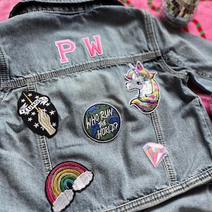 May include: A blue denim jacket with various embroidered patches, including a rainbow, a unicorn, a diamond, a globe with the text "Who Run the World?" and a hand holding a wand with stars. The jacket also has the initials "PW" embroidered on the back.