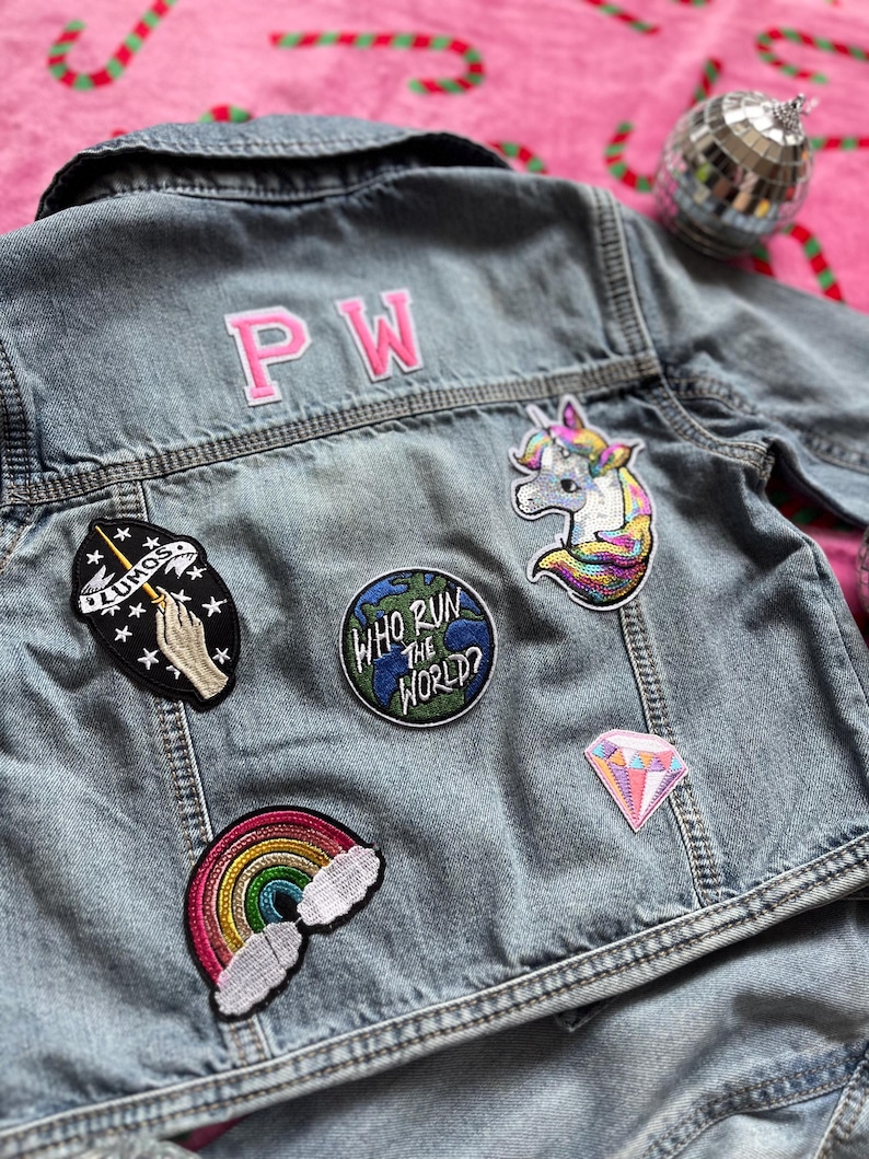 May include: A blue denim jacket with various embroidered patches, including a rainbow, a unicorn, a diamond, a globe with the text "Who Run the World?" and a hand holding a wand with stars. The jacket also has the initials "PW" embroidered on the back.