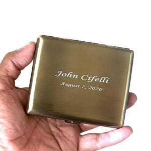 May include: A rectangular, brushed brass-toned metal case featuring a hinged lid. The surface is engraved with the name John Cifelli and the date August 7, 2026, in a clean, white-toned script and serif font style across the center.