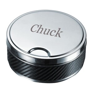 May include: A round, silver-colored ashtray with a carbon fiber pattern on the side. The lid is engraved with the name "Chuck" in a simple font. The ashtray has a small notch on the side for resting items.